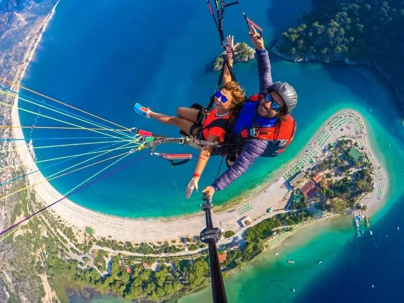 A tandem paraglide seen from above, with a beach and turquoise and blue sesn beneath