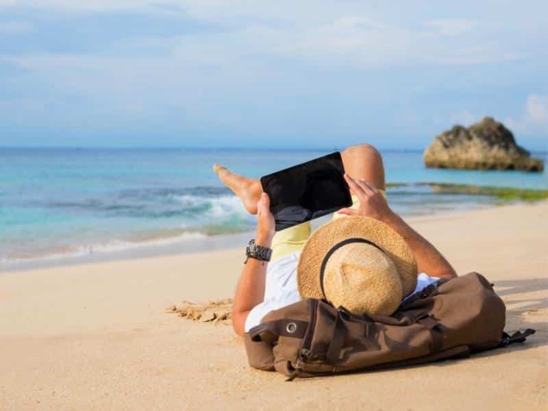 man lying on a sandy beach resting his head on a travel bag, wearing a straw hat and looking at an iPad.