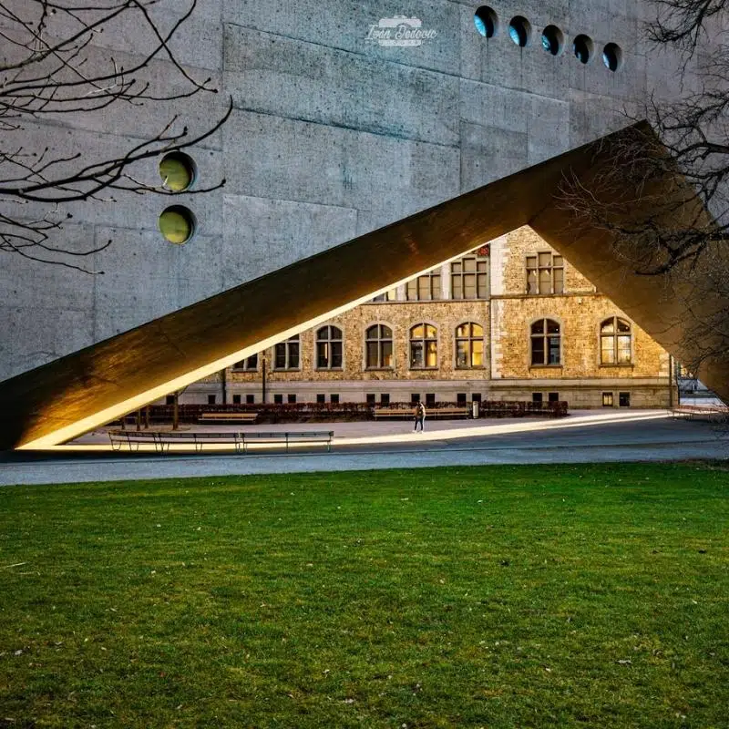 An image of a v shaped concrete building, through the bottom of the v you see parts of an oder building bathed in sunlight.