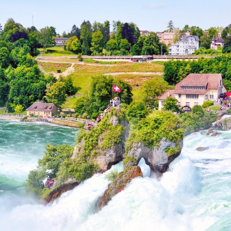 A close up image of the Rhine falls with houses and a swiss flag in the background