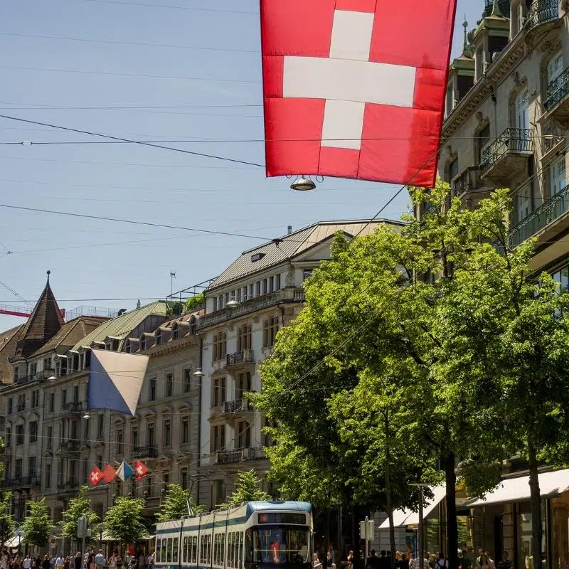 An image of a cable car on a busy shopping street , with trees and a Swiss flag in the forefront.