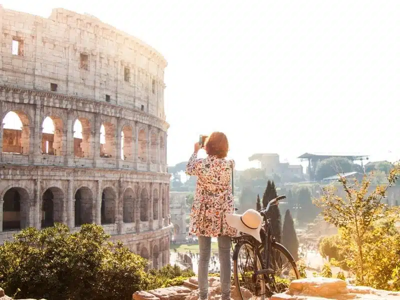 Woman wearing a flowery shirt taking a photo of the Colosseum in Rome as she stands by a bicycle.