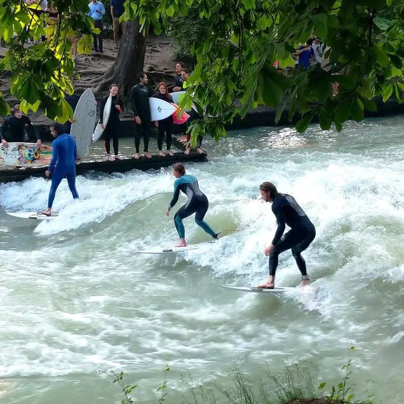 This photo shows people surfing on a river wave, surrounded by trees and onlookers waiting with their surfboards.