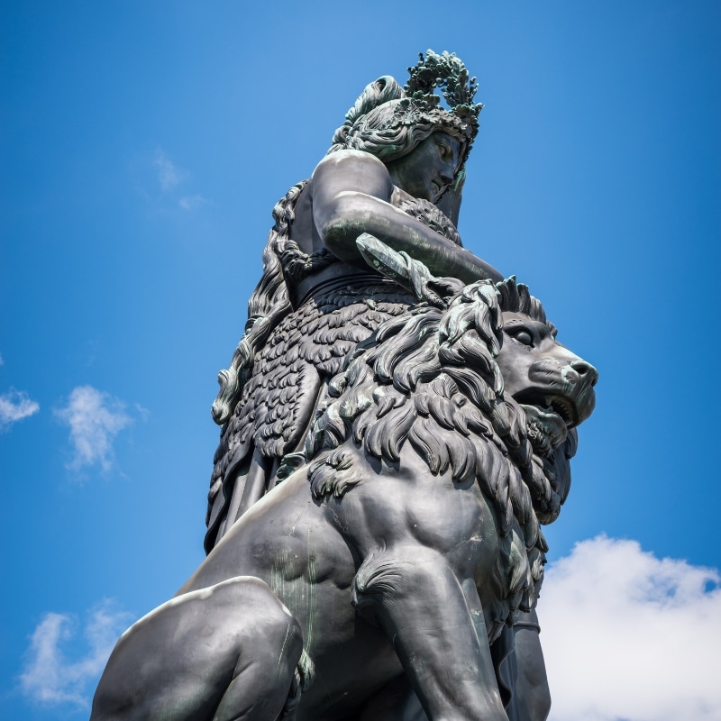 A close up image of the top of the Bavarian Statue, with a lion and women made of bronze, against a bright blue sky.





