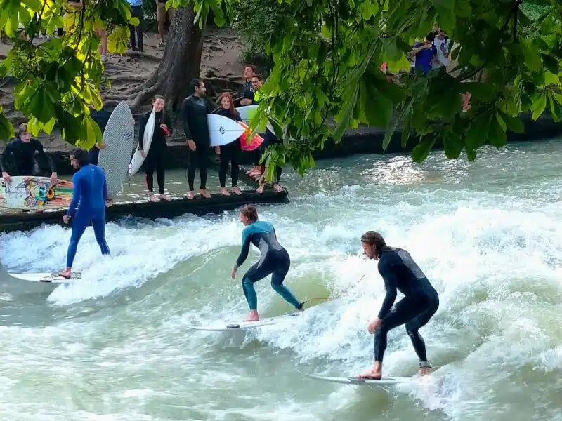 People surfing on a river wave, surrounded by trees and onlookers waiting with their surfboards.