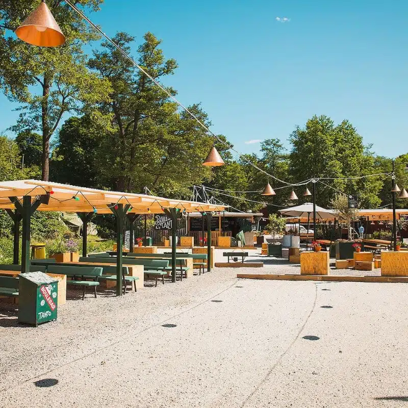 an image of a boule court surrounded by tables and benches and a bar in the background, lights are strung up for playing at night.