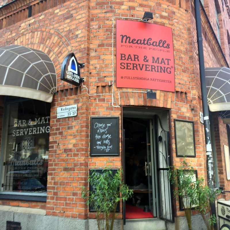 A rounded brick building on a corner, with grey awnings over the windows and a red sign with black writing saying meatballs for the people