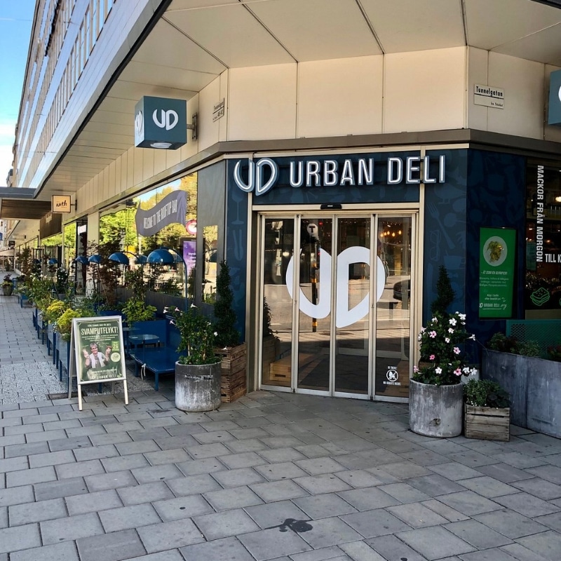 This image shows the front of a restaurant with modern glass doors and a blue surround, the sign is in neon and reads urban deli.