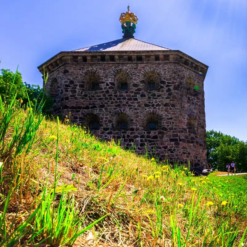 An image of an old hexagonal building on the side of a grass hill, with blue sky in the background.