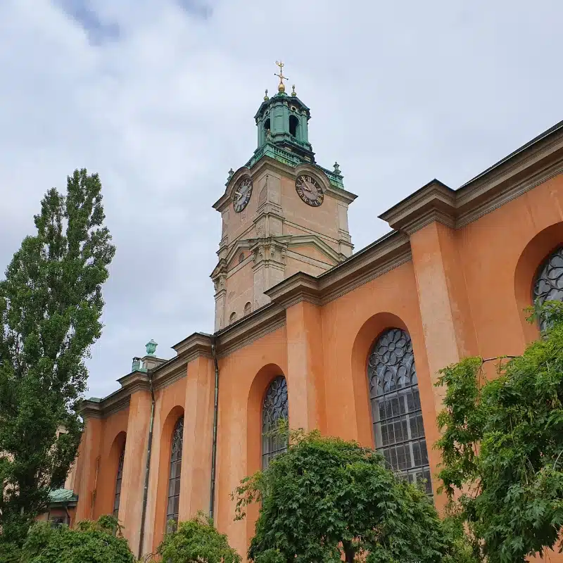 This image shows Stockholm cathedral, it has an orange facade with decorative arch windows and a clock tower with a green copper top