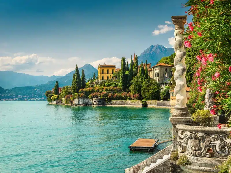 A yellow painted Italianate villa by the side of a lake, surrounded by pink oleander plants and conifer trees