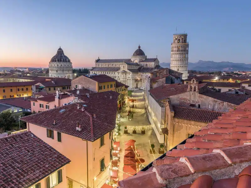 a leaning tower, domed cathedral seen beyond the roofs of a city at dusk