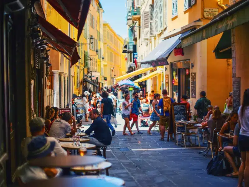 Poeple strolling and sitting on a busy French street lined with colourful houses