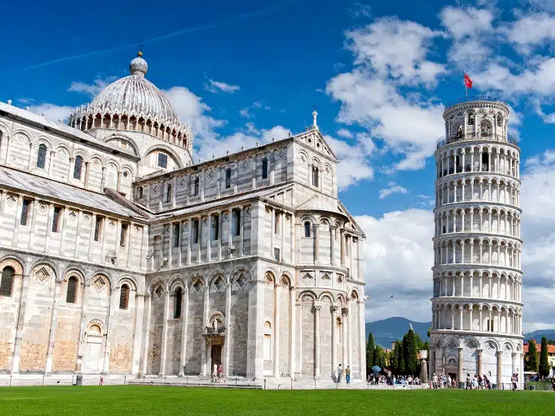 A white marble leanng tower next to a large white marble church with dome on a grass field