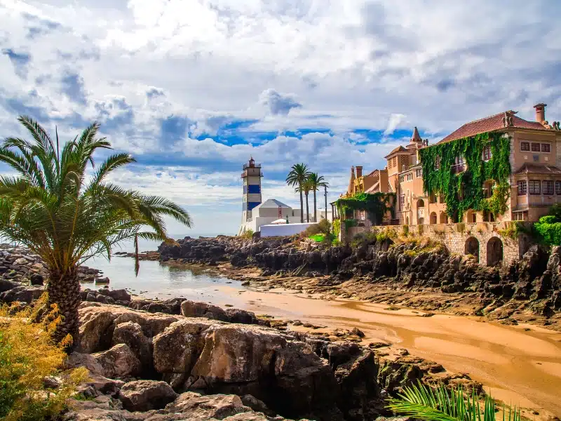 Historic building above sand with a blue and white lighthouse in the distance, and a palm in the foreground