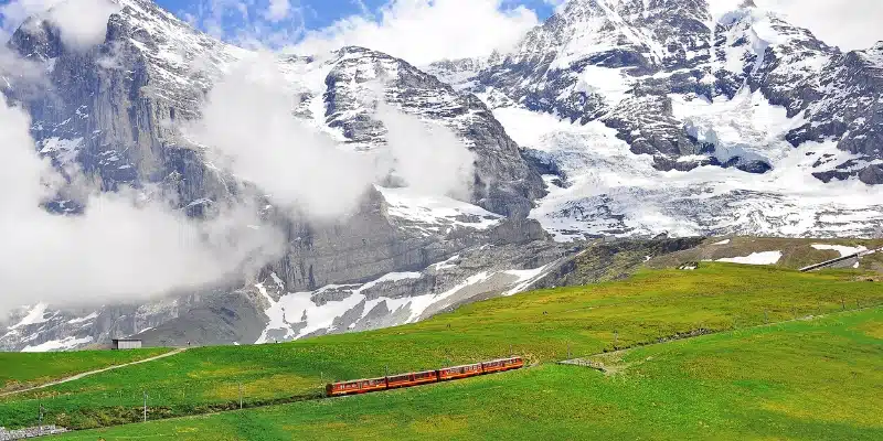 small red train in a green meadow in front of mountains and low cloud