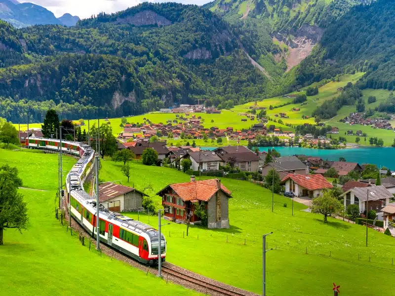 Red and white passenger train passing a small Swiss village with turquoise lake and green fields.