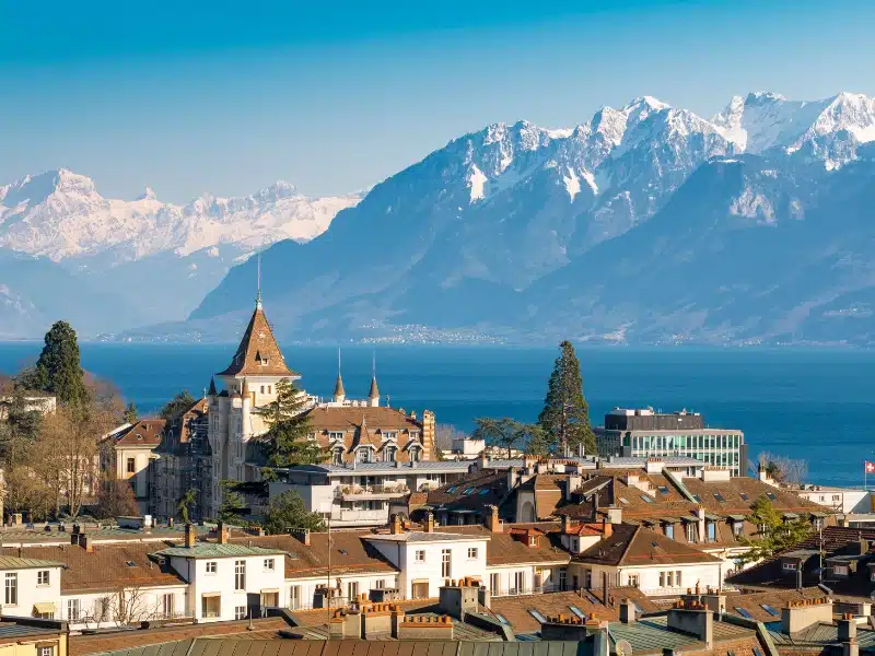 Large snow capped mountains seen across a blue lake and brown rooftops