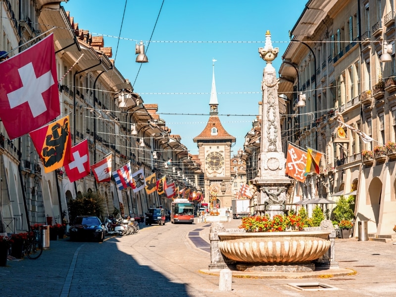 City street with hanging flags, a short and squat closk tower and a red bus