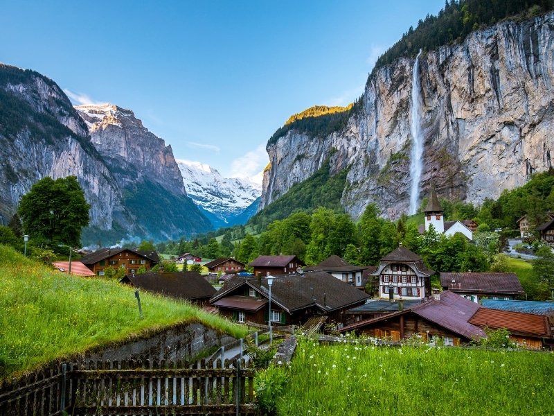View across green grass to Swiss chalets and tall mountains with a waterfall and blue sky