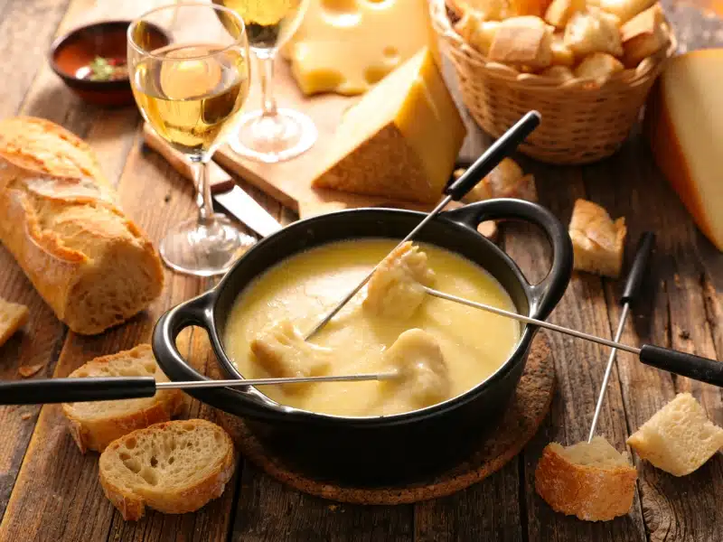 Black pot of melted cheese and skewers on a wooden table, with a glass of wine and bread on display.