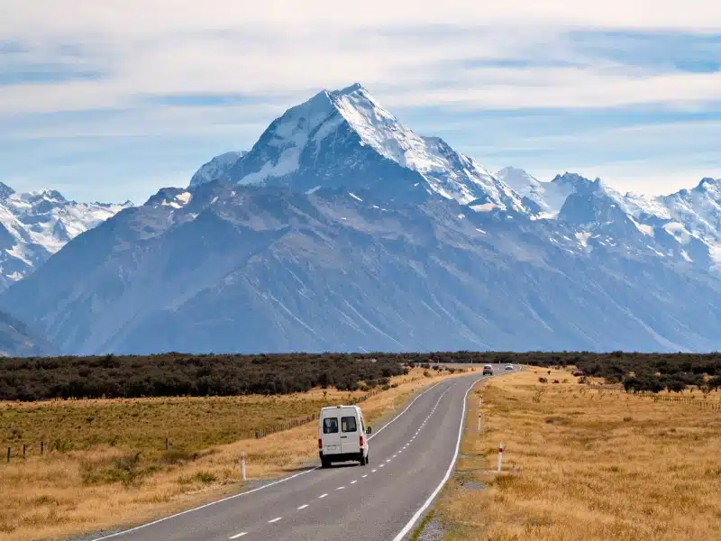 White campervan on a road with yellow grass and green vegitation either side and large snow capped mountains in the distance.