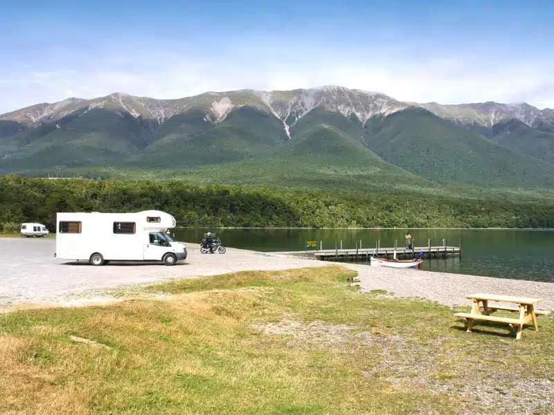 motorhome parked by a lake with a wooden jetty and nearby picnic table, with green covered mountains in the background.