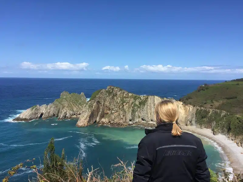 Woman with blonde hair in motorbike clothes overlooking a sandy beach with turquoise water.