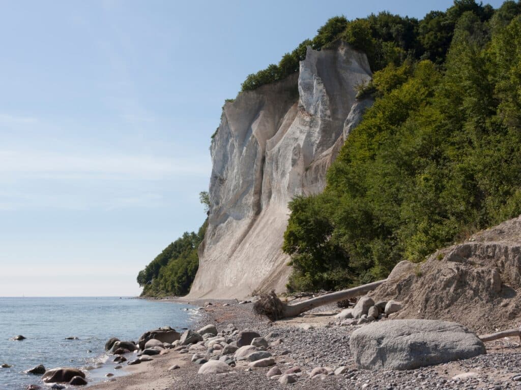 White cliffs above a narrow pebble beach and sea, with trees around the cliffs