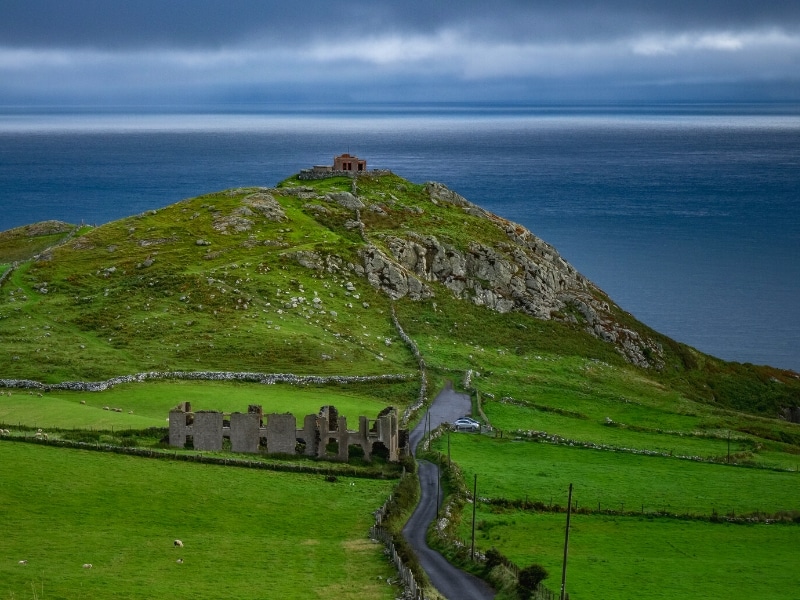 Green fields surrounding a narroe road which passes a ruin on the way to the sea