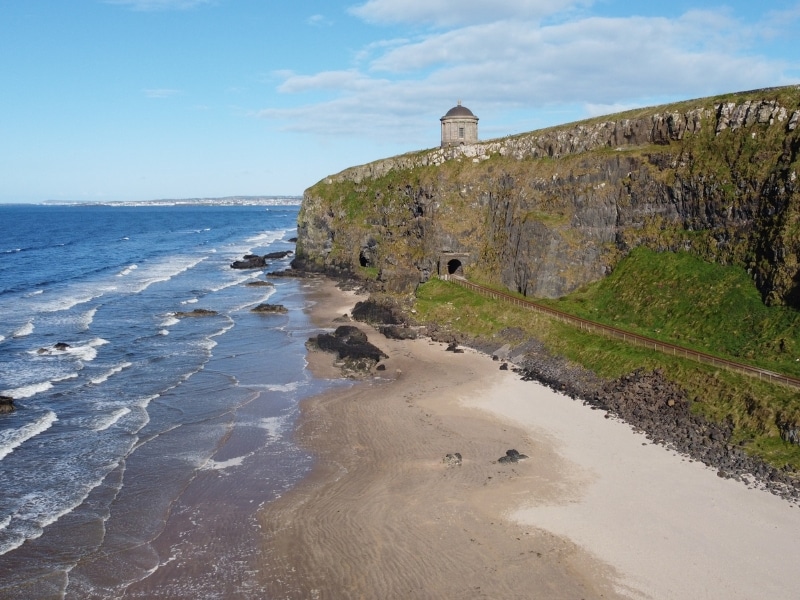 A round temple on a clifftop above a sandy beach and a railway tunnel