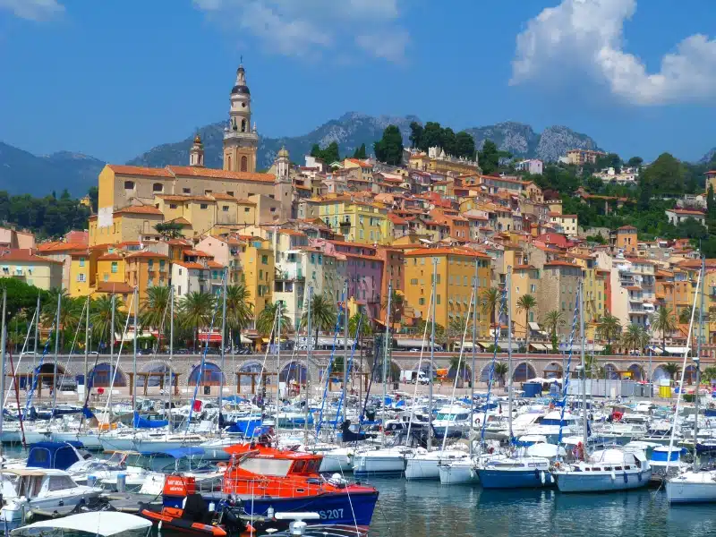 Colorful French houses behind a marina full of sailing boats, with mountains in the dustance
