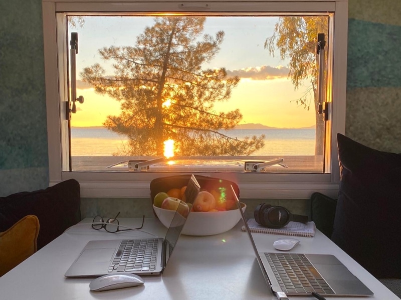 two laptops on a table in an expedition truck with a view out of the front window to the sunset, a tree and sea beyond.