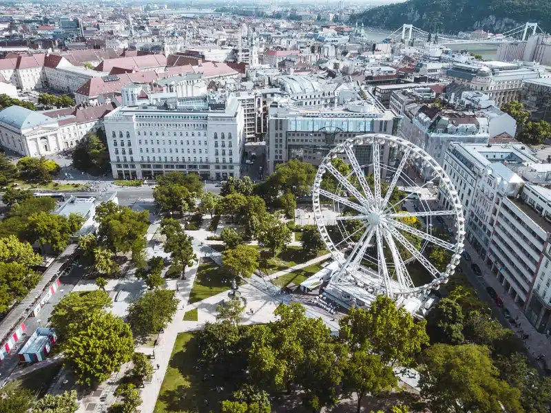 An image of a n inner city square with a large ferris wheel and lots of grass & trees