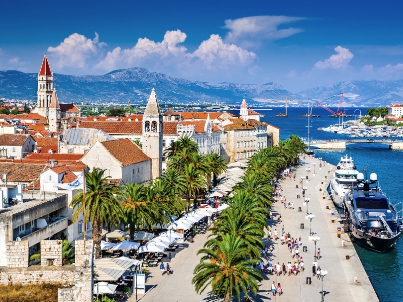 red roofed buildings and several church spires with a wide seafron promanade next to a boat filled harbor