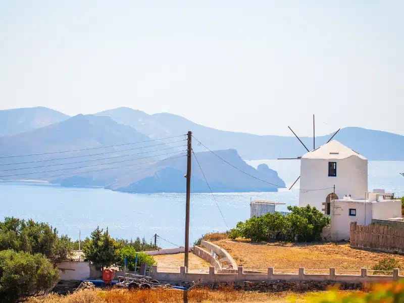 white Greek windmill with small wooden sails against a backdrop of sea and mountains