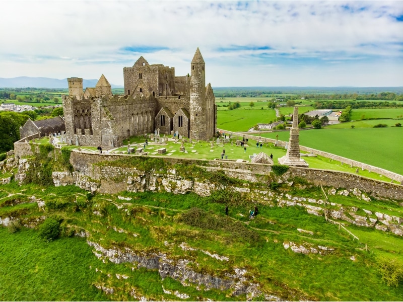 View of a group of historic buildings in a walled enclosure on a rocky outcrop surrounded by green fields