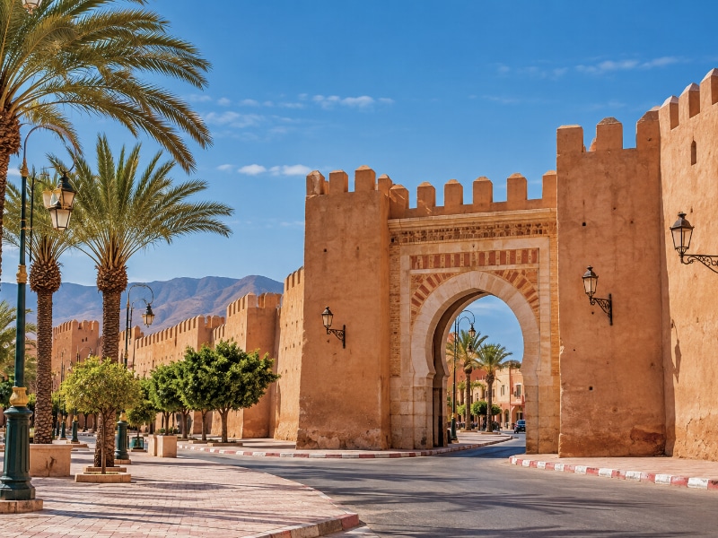 Tiznit, Morocco, showing ochre-coloured fortified walls, a traditional arched gateway and palm-lined streets beneath a clear blue sky.