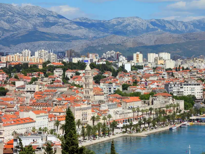 Creamy buildings in a city backed by mountains and with a seafront lined with palm trees