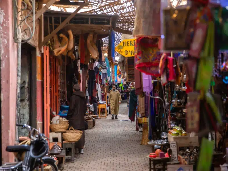 Man in a djellaba walking in a narrow street in a souk lined with colorful fabrics.