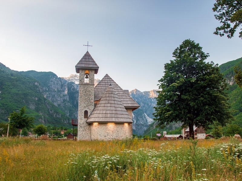 small stone chrurch with a wooden tiled roof surrouned by mountains