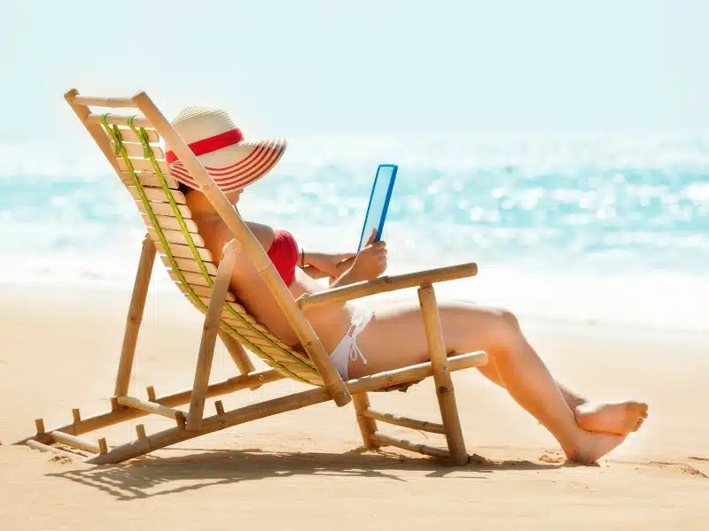 woman in a red and white bikini sitting in a deckchair on the beach and looking at a tablet
