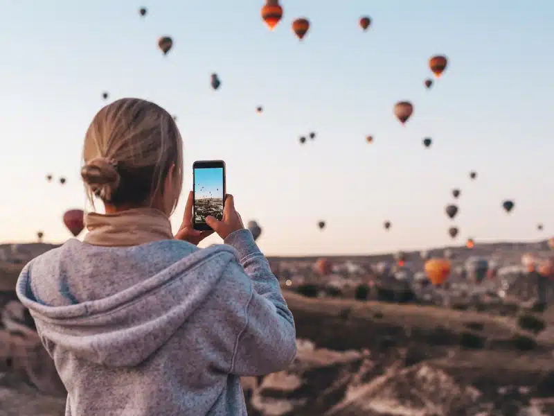 Woman with blonde hair in a bun wearing a grey hoodie taking a picture on a smartphone of multiple hot air balloons in Cappadocia, Turkey