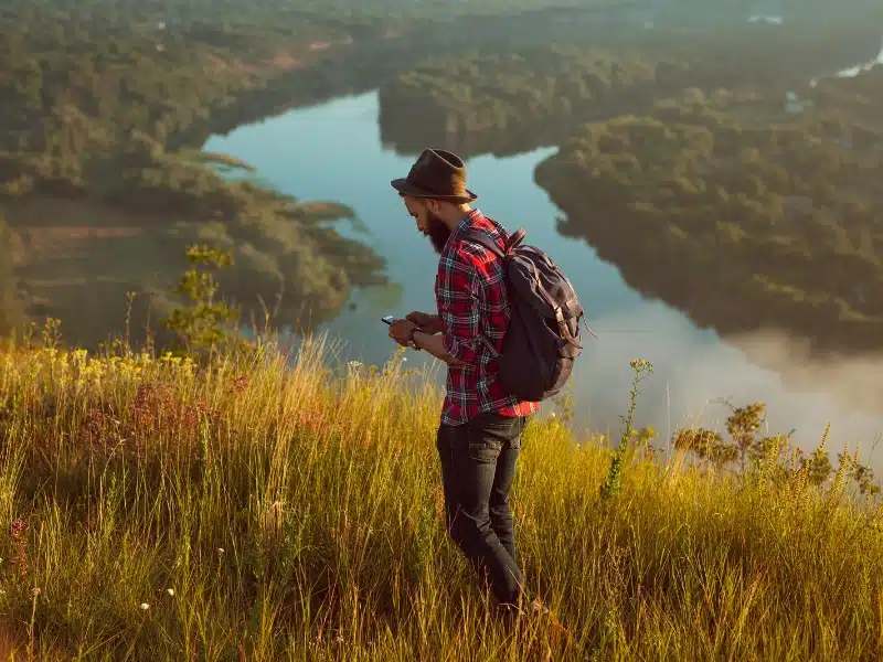Man wearing a hat and red checked shirt lookng at a phone as he stands in tall grass on a hill with a river behind him
