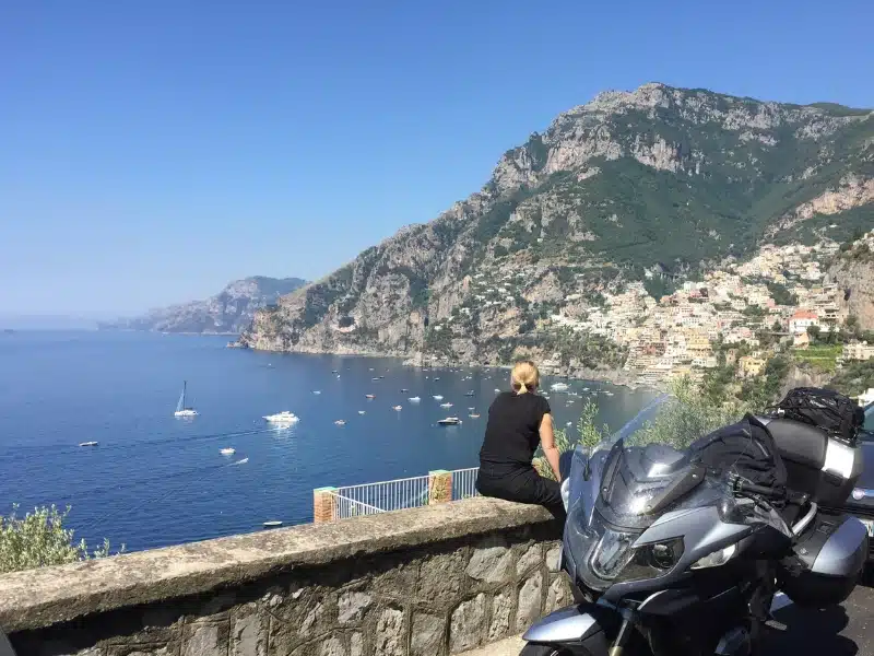 Woman sitting on a stone wall by a motorbike looking out over a blue sea and craggy mountains