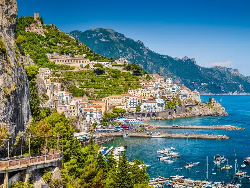 houses below a terraced green hillside facing. ablue sea, with piers and boats in the water