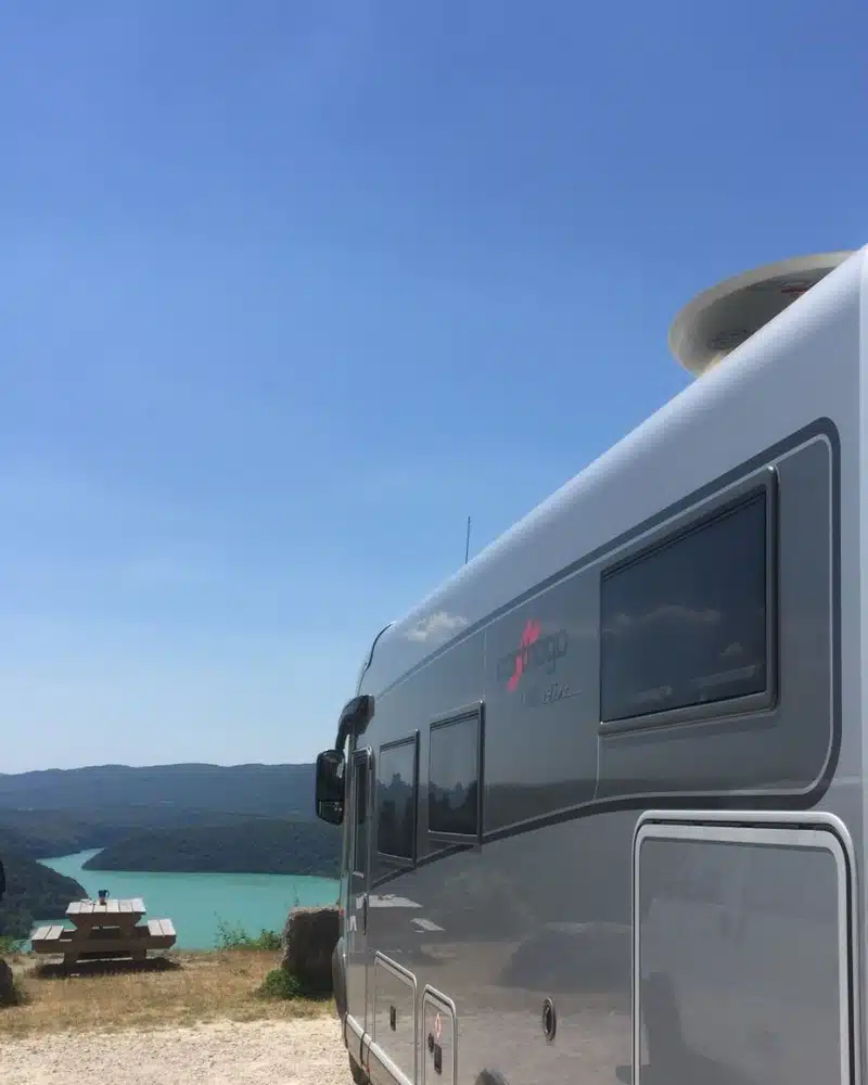 large motorhome facing a turquoise lake next to a wooden picnic table and benches