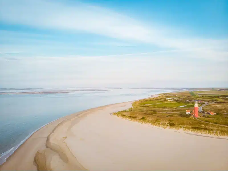 Large and deep expanse of beach with a red and white lighthouse and buildings set back in grassy sand dunes