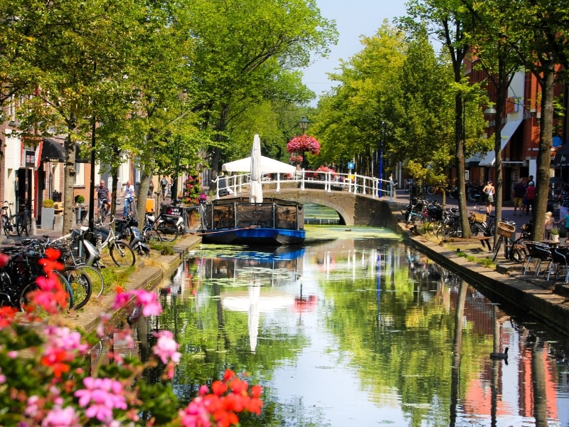 small canal lined with trees, bikes and buildings with a bridge and boat in the distance