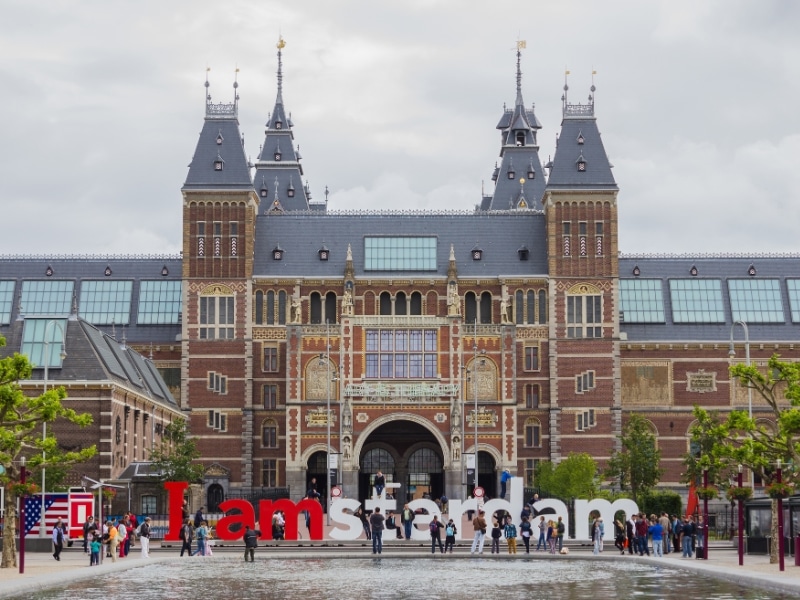 large red brick public building with a grey slate roof. the pond in front has a red and white sign behind saying 'I am Amsterdam"